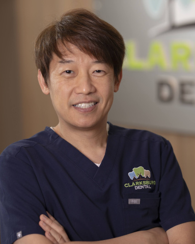 The image shows a man wearing a white lab coat, standing in front of a sign with the word  Clear  on it, posing for a professional headshot.