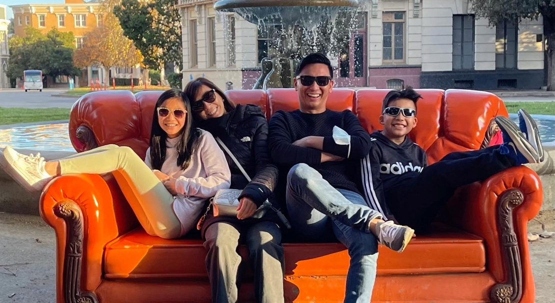 This is a photograph of three people sitting together on a red couch, with a man leaning against the backrest and two individuals seated facing him, all posing for the camera. They are outdoors, possibly in an urban setting, and there appears to be a water feature nearby. The lighting suggests it might be late afternoon or early evening.