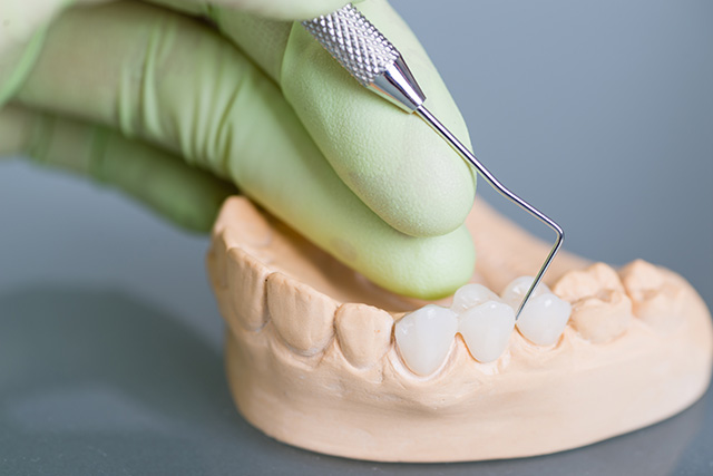 The image shows a person s hand holding a dental tool over an open mouth model with a toothpick inserted into it, indicating dental care or oral hygiene demonstration.