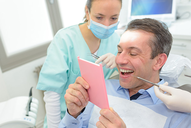 A man holding up a card while sitting in a dental chair with a smiling woman standing behind him, both in a dental office setting.