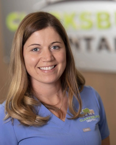 The image shows a woman with blonde hair smiling at the camera. She is wearing a blue shirt and has a name tag on her clothing. Behind her, there is a sign that reads  CROSSROADS DENTAL .