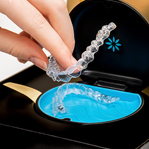 This image shows a pair of transparent dental aligners being placed into a black case with a blue interior, which has a clear lid. The person s hand is visible holding the aligner over the case.