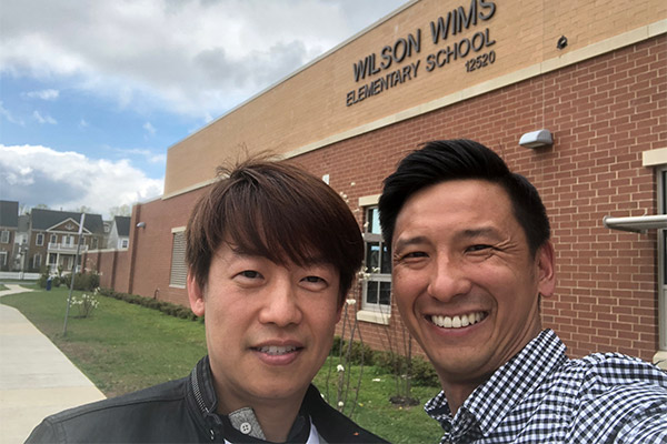 The image shows two men posing outside a school building with a sign that reads  Wilson Elementary School.  They are smiling at the camera, one man appears to be wearing a dark jacket while the other is in a lighter-colored shirt, and they both seem to be enjoying the moment.