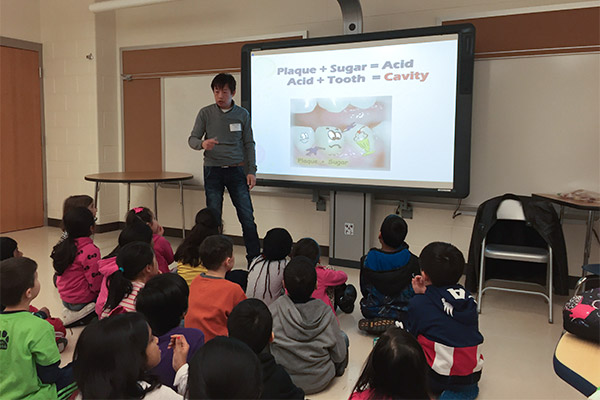The image shows a classroom setting with a teacher presenting a slide about dental health, displaying images of tooth decay and a healthy mouth, while students are seated and attentively listening.