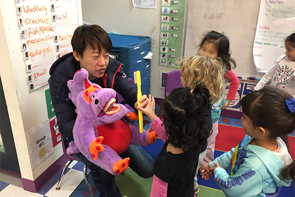 A man holding a yellow ruler while standing next to a stuffed purple dinosaur, surrounded by children in a classroom setting.