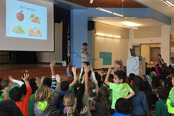 The image shows a person standing at a podium with a presentation slide behind them, addressing an audience of children sitting on the floor in front of them, who are raising their hands  the setting appears to be a classroom or auditorium.