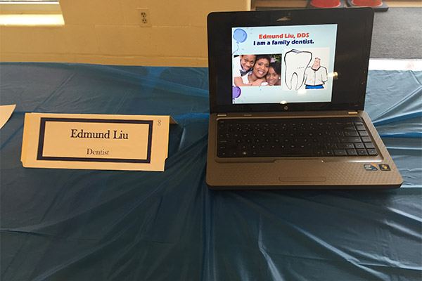 The image shows a laptop screen displaying an educational presentation with a photo of a family on it, placed on a table next to a sign that reads  Edward Li DDS  and appears to be part of a dental office setup.