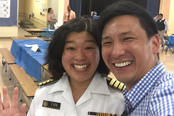 An Asian woman wearing a military uniform stands next to a smiling man at an event with tables and chairs visible in the background.