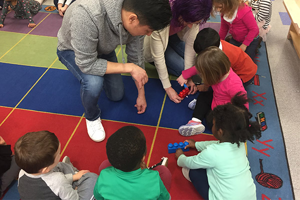 A group of people, including adults and children, are gathered around a colorful rug on the floor, with one adult kneeling down to interact with the children.
