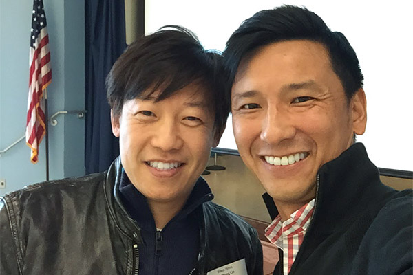 The image shows two men posing together for a photograph  one man is standing behind the other, both are smiling, and they appear to be at an event with a presentation screen visible in the background.