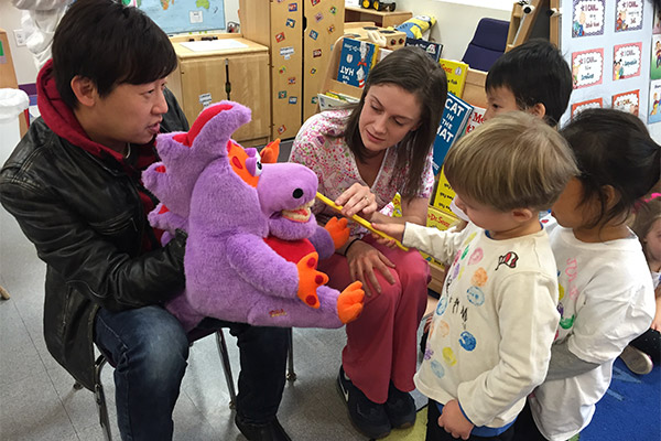 A person is holding a stuffed purple dragon toy with orange eyes, surrounded by a group of children who are looking at the toy.