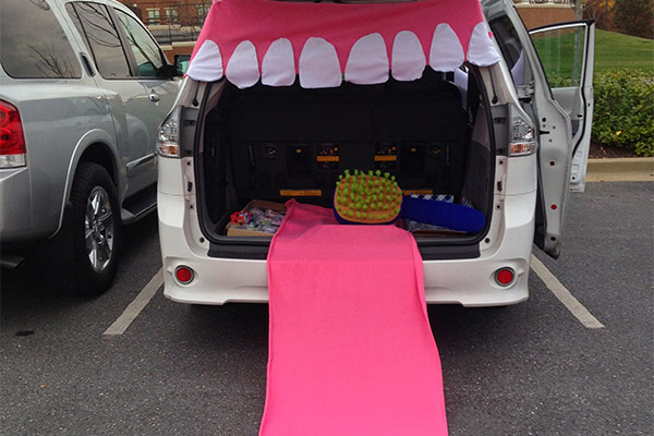 The image shows a car with an open trunk, featuring a large, pink, cartoonish mouth-shaped toy hanging from the back of the vehicle, which appears to be parked in a parking lot.