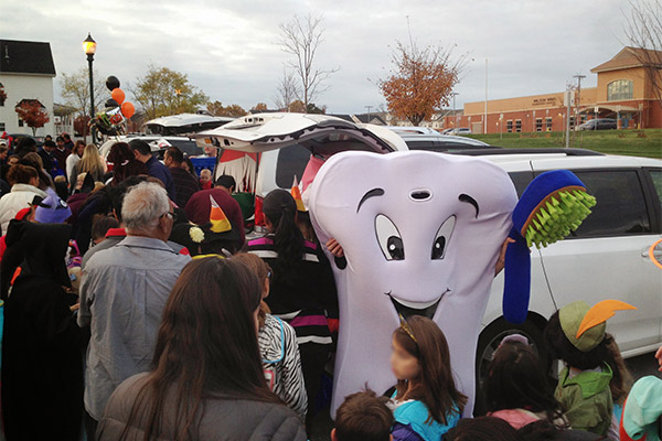 An image depicting a large, cartoonish tooth mascot wearing a white lab coat with a stethoscope around its neck, standing next to a group of people at an outdoor event during daylight hours.