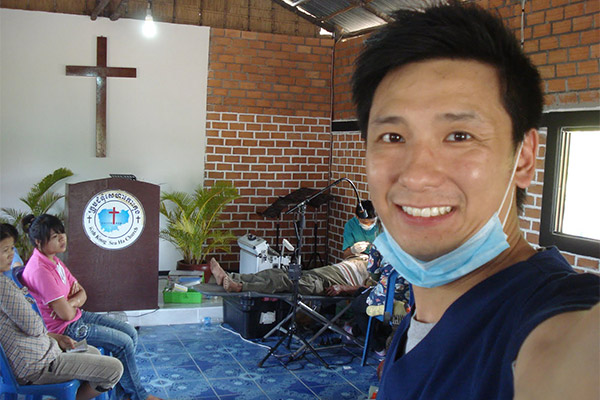 A smiling man wearing a surgical mask stands in front of a church pulpit, with a cross visible behind him.
