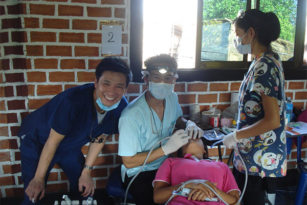 In the image, there are three people standing around a table with medical equipment, possibly in a dental or medical setting, with one person seated and receiving care from another who appears to be a medical professional.