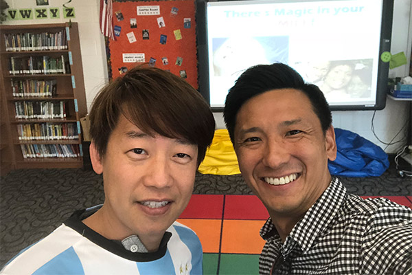 The image shows two men standing in front of a presentation screen with a slide titled 'There's magic in your MIND.' One man is smiling at the camera, while the other appears to be taking a selfie. They are indoors, and there is a classroom setting behind them.