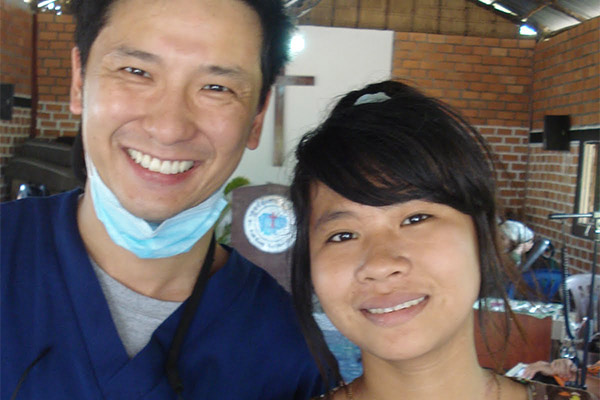 A man and woman posing together; the man is wearing scrubs and a surgical mask, standing behind a table with a microphone, while the woman is smiling at the camera.