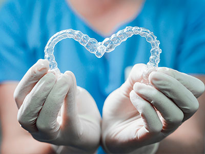 A hand holding a clear, heart-shaped acrylic retainer with a blue background.