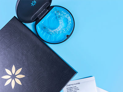 The image shows a close-up of an open package with a blue lid containing a product, placed on a surface next to a smartphone with a blue case and a clear screen protector, set against a blue background.