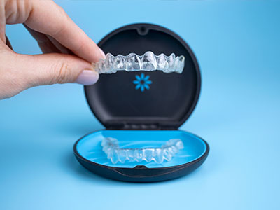 The image shows a hand holding a clear plastic tray containing a set of transparent dental aligners, with one aligner being removed from its packaging and placed on top of another aligner inside the case, which is open and resting on a blue surface.