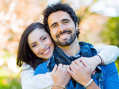 A man and woman hugging each other with smiles on their faces, set against a blurred outdoor background.