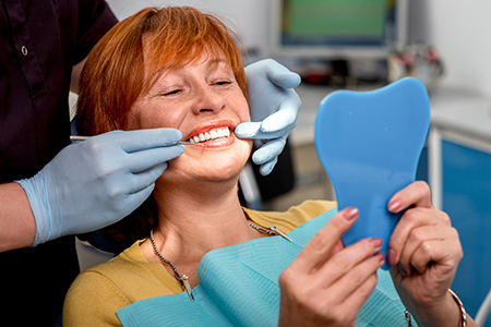 A woman in dental office holding up a blue model tooth while smiling broadly at camera.